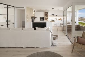 A modern living room with a white interior, featuring a large sofa, a kitchen island, and a balcony with a view of the city.