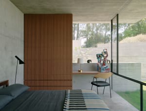 Modern minimalist bedroom featuring concrete walls and floor, with a wooden wardrobe and a glass desk with a colorful sculptu