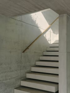 A minimalist staircase made of concrete with a wooden handrail, illuminated by natural light.