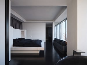 A minimalist bedroom with a white concrete bed platform, black bedding, and a large window.