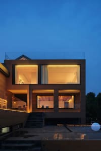 A modern, two-story house with a concrete exterior and large windows, illuminated at dusk.