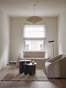 A minimalist residential interior featuring neutral textiles, metal tables, and a large window with light-colored curtains.