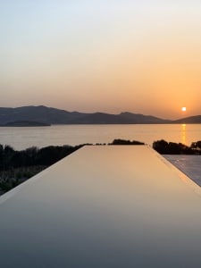 A minimalist swimming pool with an infinity edge overlooks a sunset view over the sea and mountains.