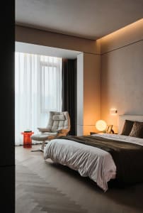 A minimalist bedroom with white and blush tones, featuring a modern armchair, a cozy bed with a textured blanket, and a warm 