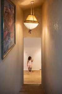 A young girl in a tutu skirt plays under a hanging lamp in a children's room with wooden stairs and walls.