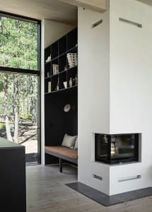 A minimalist residential interior with a concrete fireplace, glass walls, and a wooden floor and bench. Large windows provide