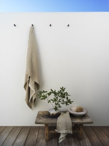 Minimalist bathroom scene with a ceramic plant on a wooden bench, a textured towel hanging on the wall, and a wood and cerami