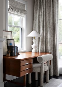A modern home office featuring a wooden desk, fabric-upholstered chair, and glass-paneled window with patterned curtains.