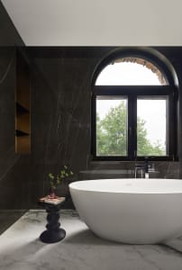 A modern bathroom featuring a dark marble wall, a white freestanding bathtub, and an arched window overlooking green trees.