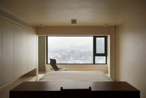 A minimalist bedroom with a large window overlooking a snowy cityscape, featuring a bed, a chair, and a desk.