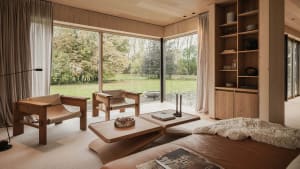 Modern living room with wooden furniture, large windows showing greenery, and a cozy atmosphere.