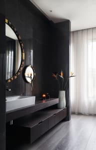 A modern bathroom featuring black marble walls, a floating vanity with a white sink, and a vase of flowers by the window.