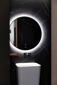 Modern bathroom with a sleek, round mirror with backlit LED lighting and a white ceramic pedestal sink against a black marble