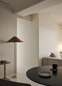 A minimalist living room with a neutral color palette, featuring a sofa, a table with a plate and mug, and a lamp with a coni