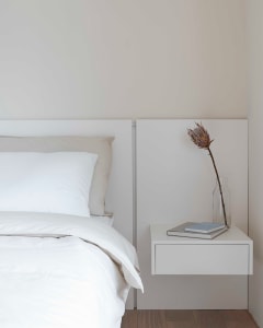 A serene, minimalist bedroom featuring a white bed with neatly arranged white bedding and a simple, white nightstand with a v