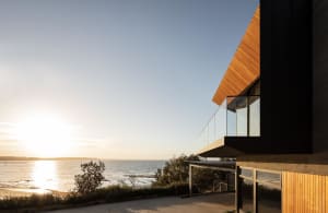 A modern seaside residential property featuring wooden cladding and large glass windows overlooking the ocean.