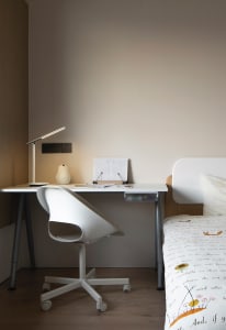 A modern minimalist bedroom with a white desk and chair, a bed with a white and floral comforter, and a white desk lamp.