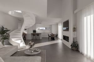 A modern living room featuring a white stone floor, a spiral staircase, a round coffee table, a minimalist sofa, and a kitche