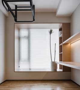 A modern minimalist home office featuring a wooden floor, blinds, potted plants, and a shelf.