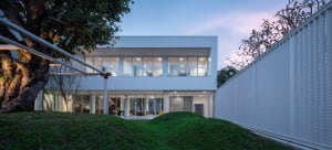 A modern house with large glass windows and a concrete facade viewed from the backyard at dusk.