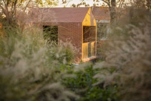 A minimalist brick house with a small window and a simple roofline surrounded by tall grasses and trees.