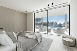Modern minimalist bedroom with glass walls overlooking a serene outdoor space with a pool and lounge areas.