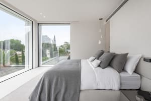 A modern, brightly lit bedroom with a bed dressed in light fabrics and glass windows.