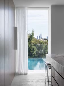 A modern residential kitchen features a glass window overlooking an infinity pool and city skyline, with marble countertops a