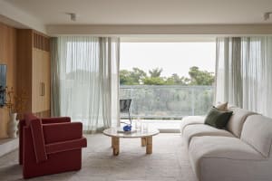 A modern living room with neutral tones and a view of greenery outside through sheer curtains and a glass barrier.