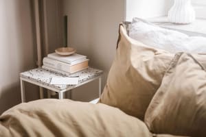 A modern bedroom featuring white textured cotton sheets on a bed, a small side table with books and a candle holder, and a wi
