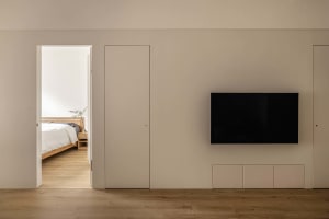 A minimalist apartment with a living room featuring a wooden floor, a television mounted on a wall, and access to a bedroom w