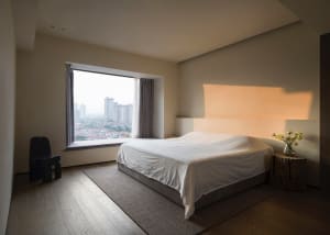 A minimalist bedroom with a large window providing a view of the cityscape, featuring a bed with a white sheet and a wooden s