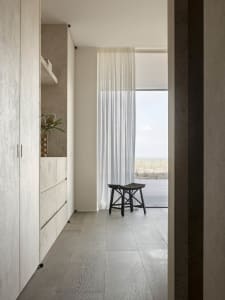 A minimalist interior featuring a light wood floor, large windows with sheer curtains, and a stool on a wooden deck overlooki