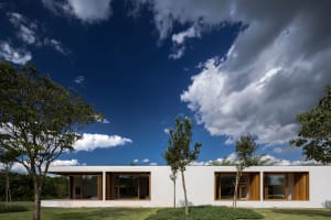 A modern residential building with white plaster exterior, large windows, and a flat roof surrounded by trees and greenery ag