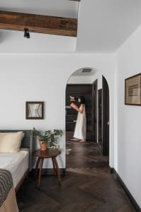 A minimalist bedroom featuring dark wooden flooring, a bed with neutral bedding, a side table with a plant, and a woman stand
