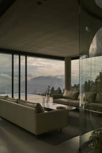A modern living room with panoramic views of mountains and a cityscape through large glass windows, featuring concrete walls 