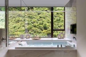 Modern bathroom with white marble walls, a large window overlooking a lush green forest, and a soaking tub with jets.