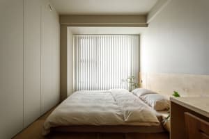 A minimalist bedroom featuring a simple wooden bed with a light-colored headboard and white bedding, positioned against a wal
