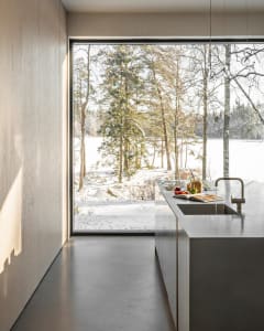 Modern kitchen with minimalist design featuring concrete countertops and floor, surrounded by a snowy forest view through lar