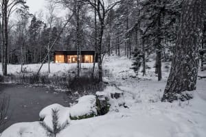 Modern residential cabin surrounded by snow-covered trees and a frozen pond at dusk, illuminated from within.