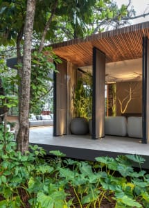 Modern living room with concrete walls, lighting, and white furnishings surrounded by green foliage.