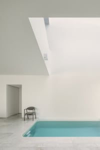 A minimalist indoor swimming pool with a white marble floor, surrounded by white walls and a ceiling with a skylight.