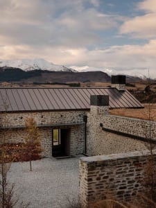 A stone cottage with a metal roof situated on a gravel driveway with a backdrop of snow-capped mountains.