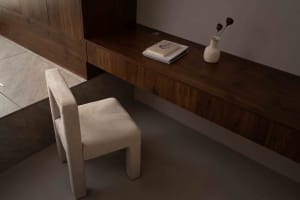 A minimalist home office desk made of wood with a stack of books and a vase on top, and a beige chair.