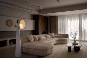 A modern living room with a curved sofa, floor lamp, and large windows with white curtains.