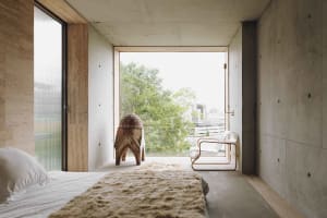 Minimalist bedroom featuring concrete walls and floor with a white bed, rug, and chair, overlooking greenery and cityscape th