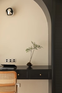 A minimalist workspace featuring a black desk with a small plant, a wooden rattan chair, and a laptop.