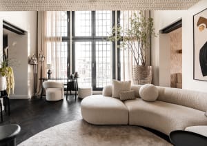 Modern living room featuring a curved, beige fabric sofa, dark wood flooring, and large windows with white curtains.