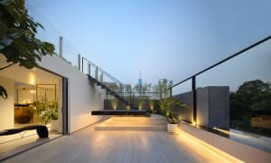 A modern rooftop garden extension with wooden floors, concrete walls, and glass railings illuminated by warm lighting.