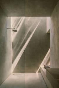 Modern bathroom with concrete walls and floor, illuminated by natural light through a window, featuring a shower head and ben
